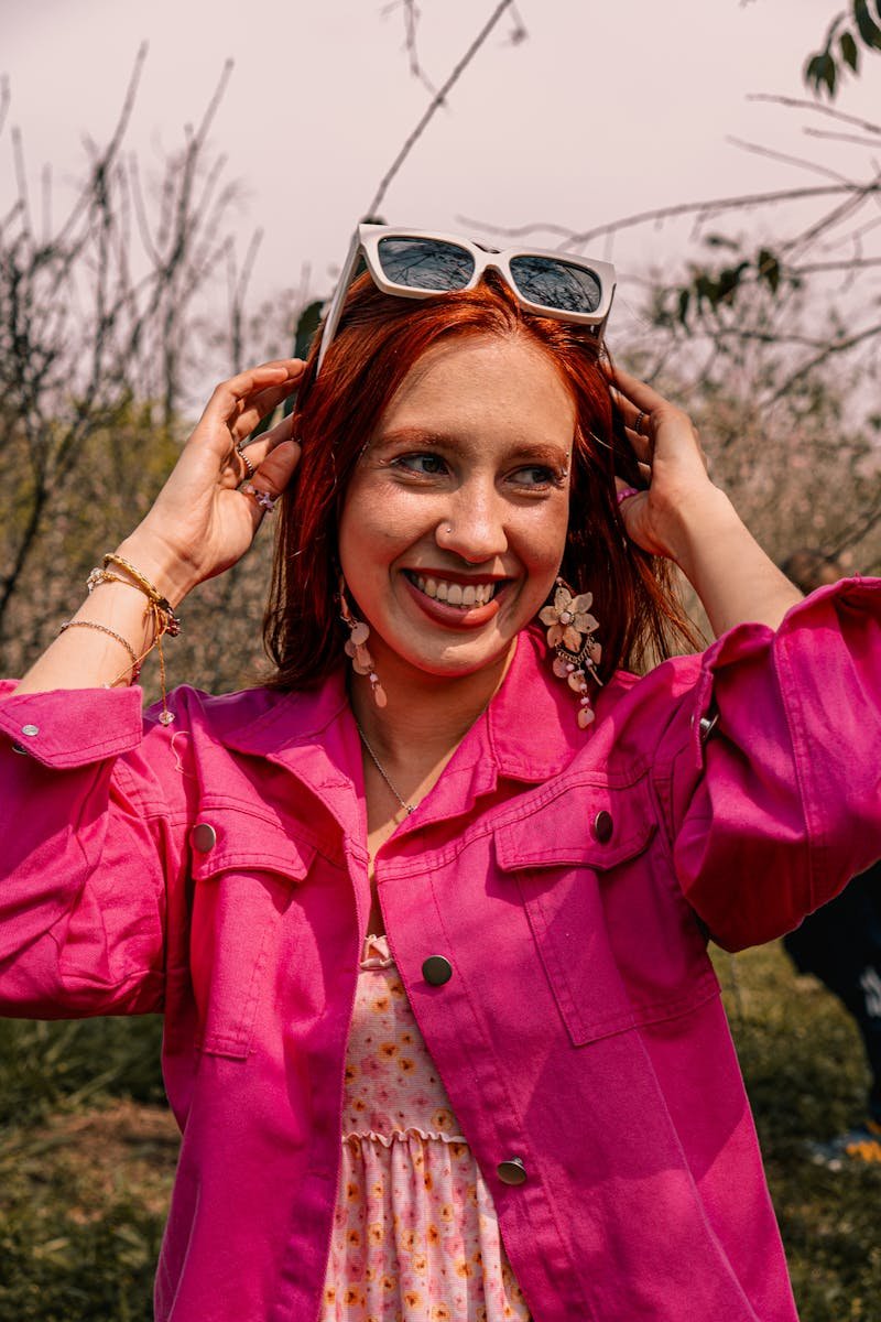 Smiling woman in a vibrant pink jacket enjoying a sunny day outdoors with stylish accessories.